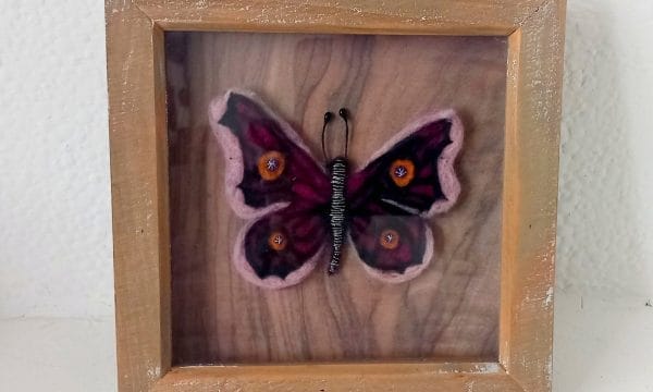 A pink and yellow butterfly made from needle felting. It has an intricate pattern on it's wings, and it's body is black with white stripes. It is in the centre of a wooden frame.