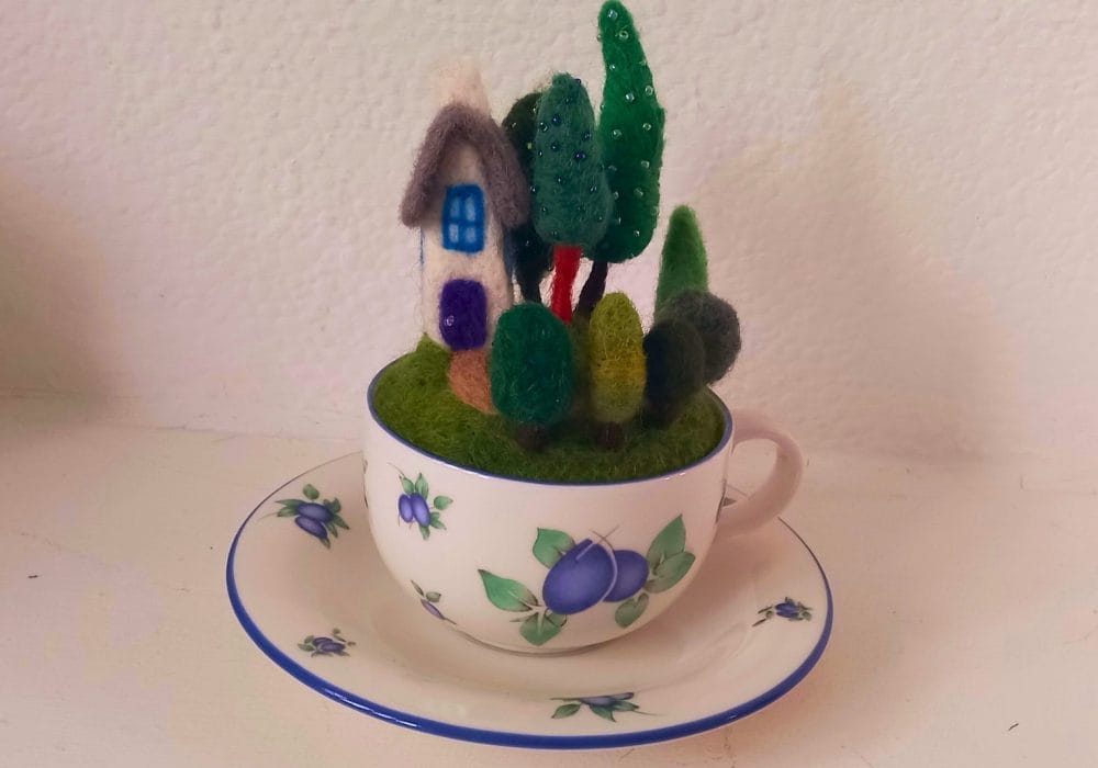 A needle felted house and garden full of green felt trees. The trees are decorated with green beads. The garden is inside of a blue and white floral teacup, sat on a matching saucer.