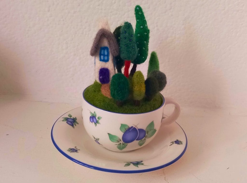 A needle felted house and garden full of green felt trees. The trees are decorated with green beads. The garden is inside of a blue and white floral teacup, sat on a matching saucer.