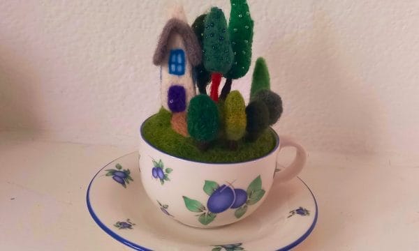 A needle felted house and garden full of green felt trees. The trees are decorated with green beads. The garden is inside of a blue and white floral teacup, sat on a matching saucer.