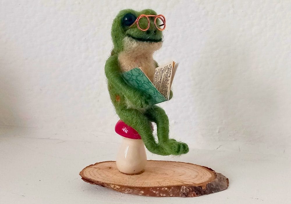 A close up of a green toad made from needle felt, it is wearing copper wire glasses, and holding a small book. It has a happy expression. It is sitting on a red toadstool.