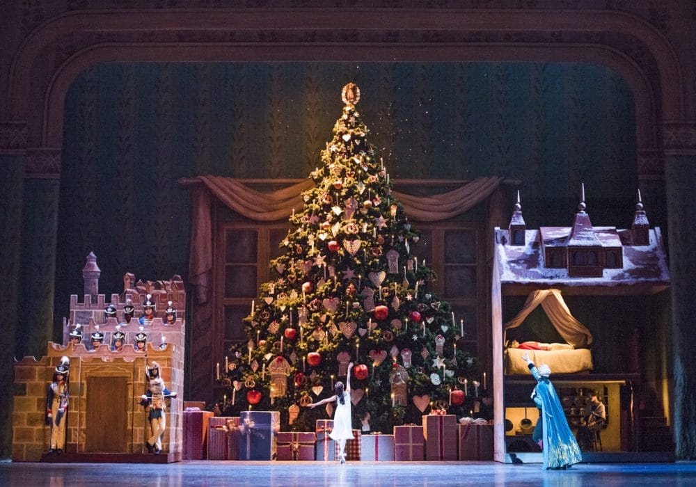 A huge Christmas tree is in the centre of a theatre stage, it is covered with red and gold decorations. To the left of the tree is a castle, with nutcrackers standing guard. To the right is a dollshouse. In front of the tree, a single ballerina stands en pointe with her arms outstretched.