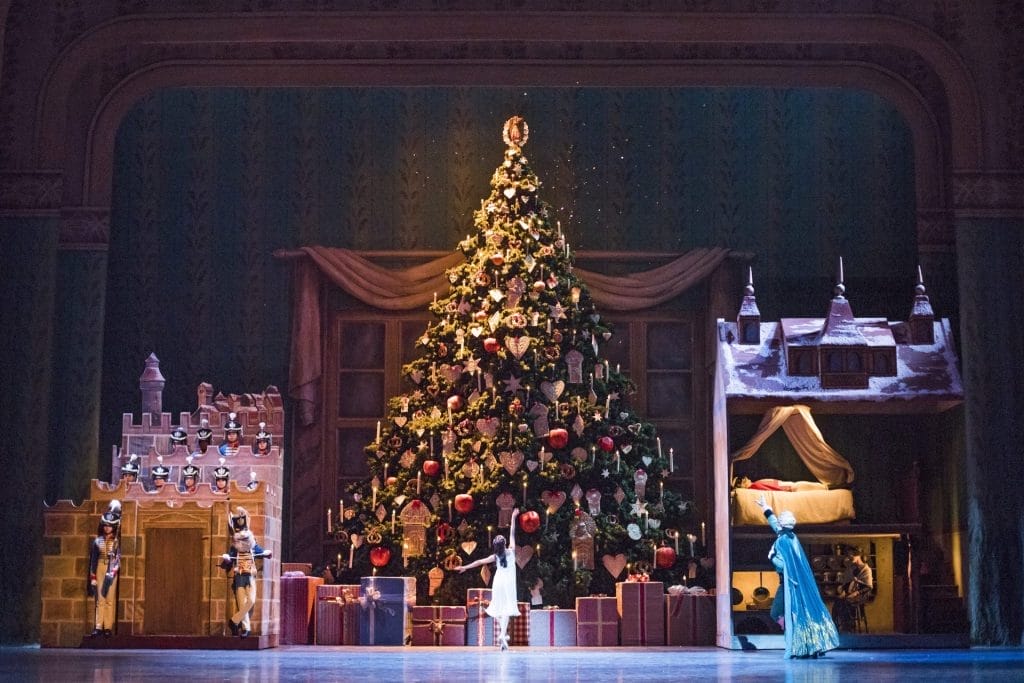 A huge Christmas tree is in the centre of a theatre stage, it is covered with red and gold decorations. To the left of the tree is a castle, with nutcrackers standing guard. To the right is a dollshouse. In front of the tree, a single ballerina stands en pointe with her arms outstretched.