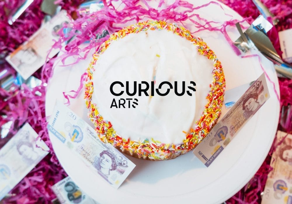 Curious Arts logo features on a cake with colourful sprinkles around the edge. The cake is surrounded by British pound notes (showing the Queen's portrait) and sits on a white plate against a background of bright pink shredded paper or confetti.