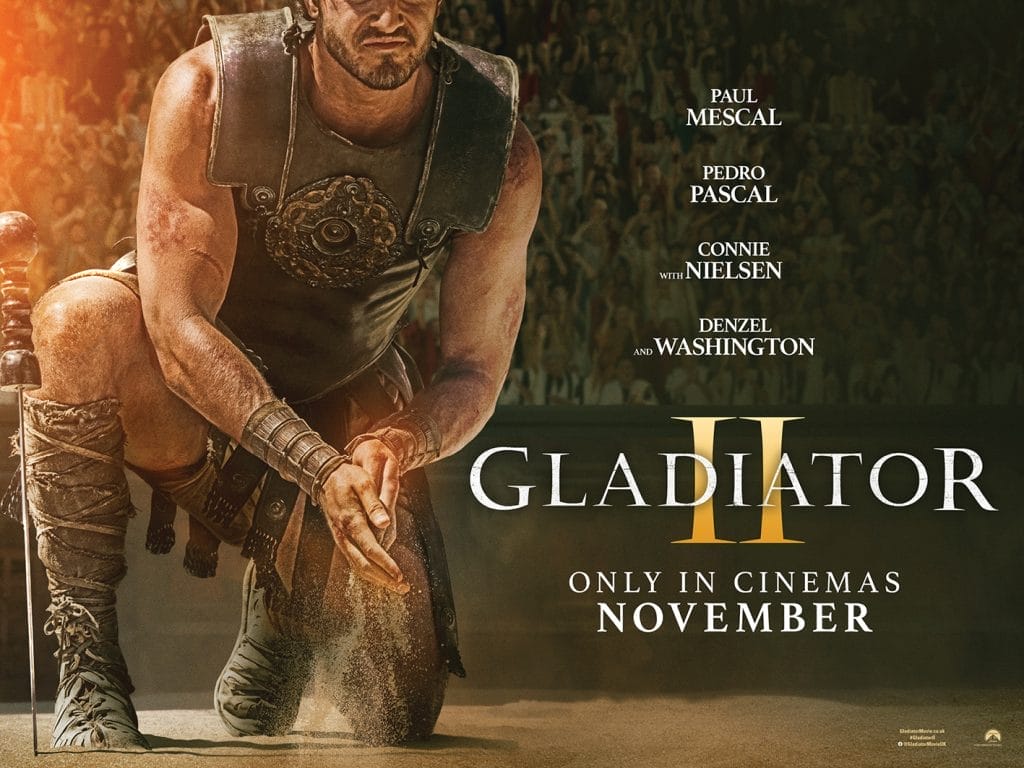 A roman gladiator kneels on sand, he is rubbing sand between his hands, his face is not visible. To the right of him is the title Gladiator II