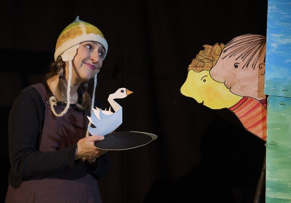 An actress wearing a colourful knitted hat is holding a paper swan puppet, she is smiling towards two paper people who are looking at the swan.