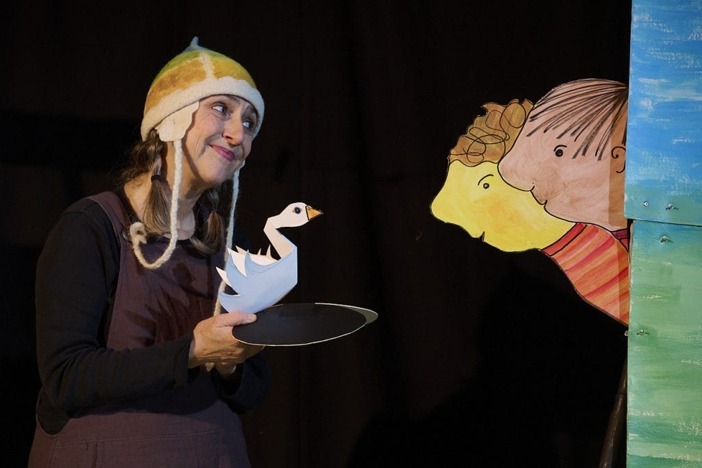 An actress wearing a colourful knitted hat is holding a paper swan puppet, she is smiling towards two paper people who are looking at the swan.