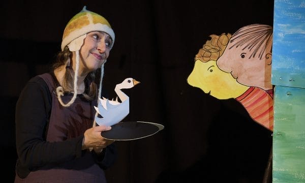 An actress wearing a colourful knitted hat is holding a paper swan puppet, she is smiling towards two paper people who are looking at the swan.