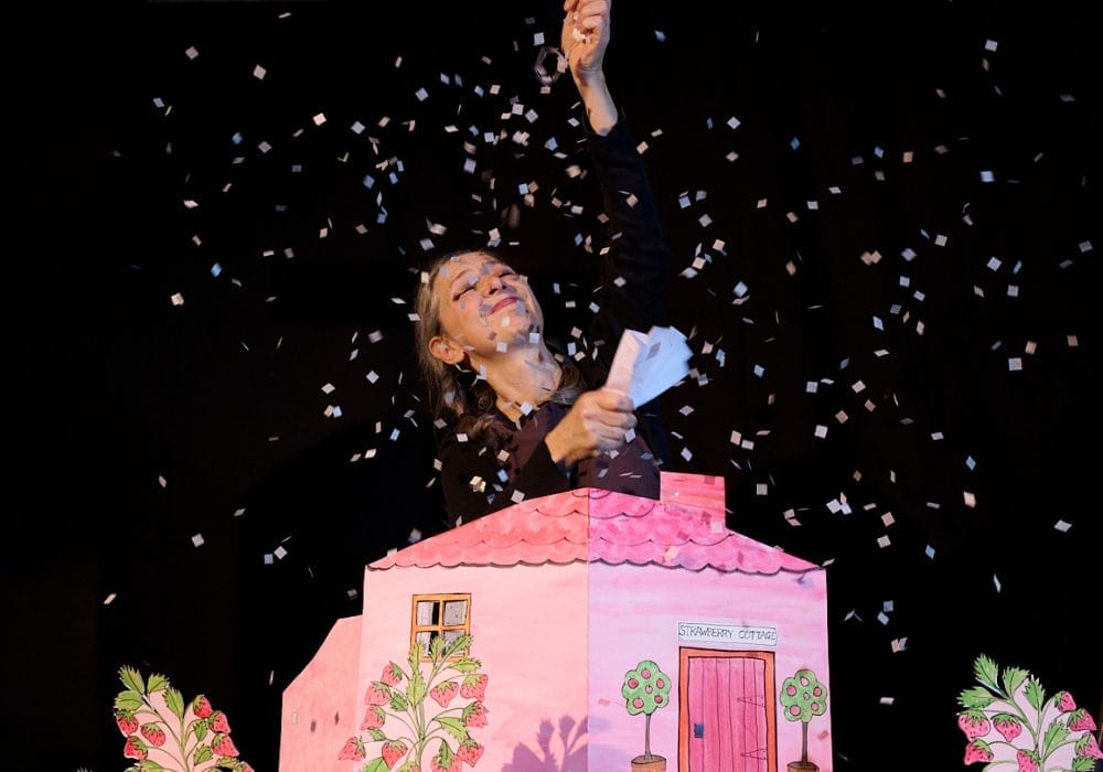 An actress is smiling with her hand raised in the air, she is surrounded by white confetti that is floating down onto a pink paper house.