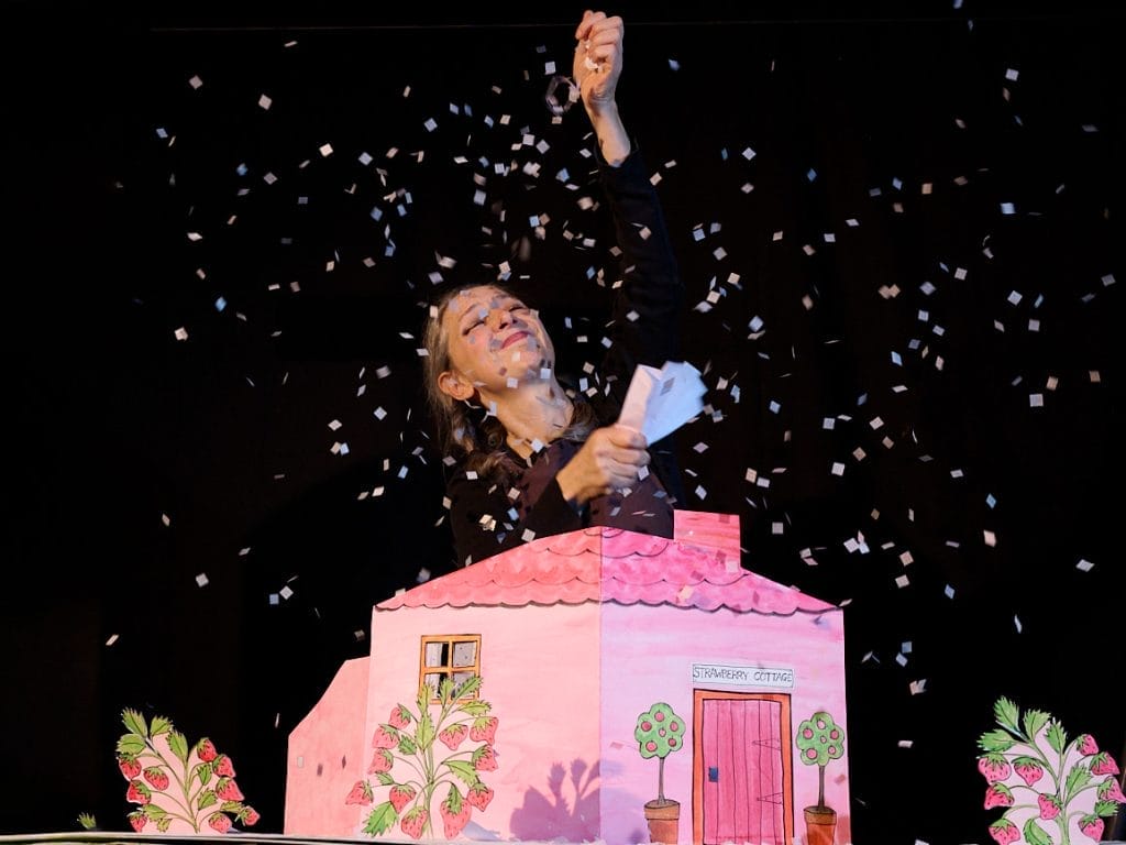 An actress is smiling with her hand raised in the air, she is surrounded by white confetti that is floating down onto a pink paper house.