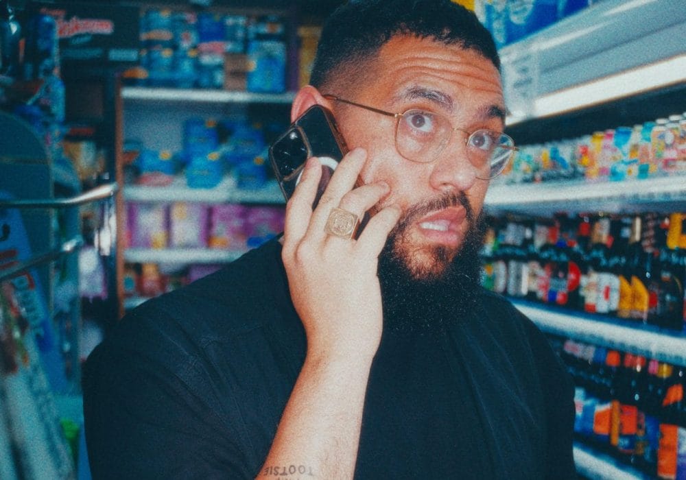 Jamali in what appears to be a convenience store or small shop. The person is wearing a black shirt and has glasses and a beard. They're talking on a phone while holding what looks like beverage bottles in their other hand. They have visible tattoos on their arm and are wearing a ring. The photo appears to be taken with a vintage or filtered effect, giving it a slightly blue-tinted color palette. The background shows store shelves stocked with various drinks and products.