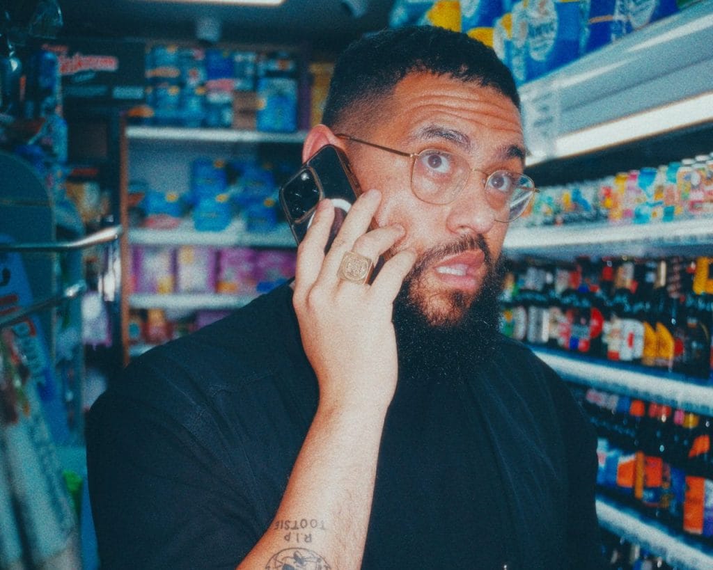 Jamali in what appears to be a convenience store or small shop. The person is wearing a black shirt and has glasses and a beard. They're talking on a phone while holding what looks like beverage bottles in their other hand. They have visible tattoos on their arm and are wearing a ring. The photo appears to be taken with a vintage or filtered effect, giving it a slightly blue-tinted color palette. The background shows store shelves stocked with various drinks and products.