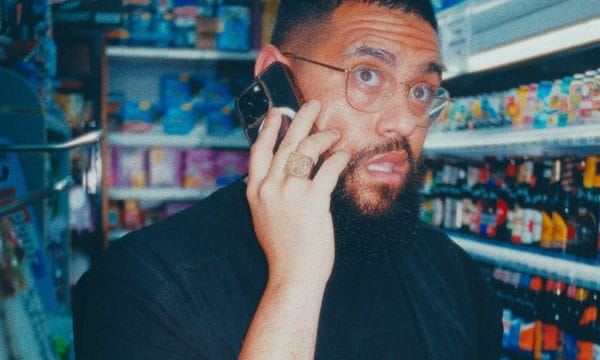 Jamali in what appears to be a convenience store or small shop. The person is wearing a black shirt and has glasses and a beard. They're talking on a phone while holding what looks like beverage bottles in their other hand. They have visible tattoos on their arm and are wearing a ring. The photo appears to be taken with a vintage or filtered effect, giving it a slightly blue-tinted color palette. The background shows store shelves stocked with various drinks and products.