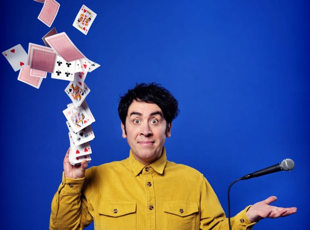 Comedian Pete Firman is looking directly into the camera, he is smiling with his eyebrows raised. A deck of cards is flying into the air from his left hand, and a microphone is floating above his right hand.