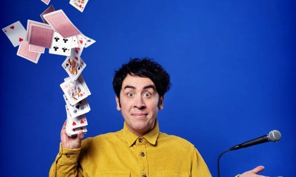 Comedian Pete Firman is looking directly into the camera, he is smiling with his eyebrows raised. A deck of cards is flying into the air from his left hand, and a microphone is floating above his right hand.