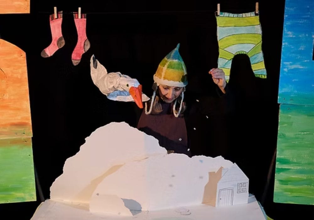 Performer Zannie Fraser is in the centre of this image, she sits behind a snowy landscape, above her is a washing line with socks and a jumper hanging from it. In one hand she holds a goose puppet, her other hand is sprinkling snow down onto the scene.