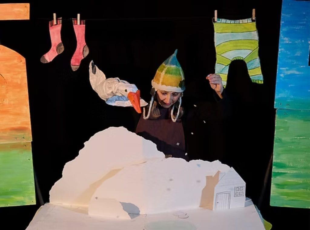 Performer Zannie Fraser is in the centre of this image, she sits behind a snowy landscape, above her is a washing line with socks and a jumper hanging from it. In one hand she holds a goose puppet, her other hand is sprinkling snow down onto the scene.