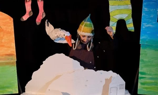 Performer Zannie Fraser is in the centre of this image, she sits behind a snowy landscape, above her is a washing line with socks and a jumper hanging from it. In one hand she holds a goose puppet, her other hand is sprinkling snow down onto the scene.