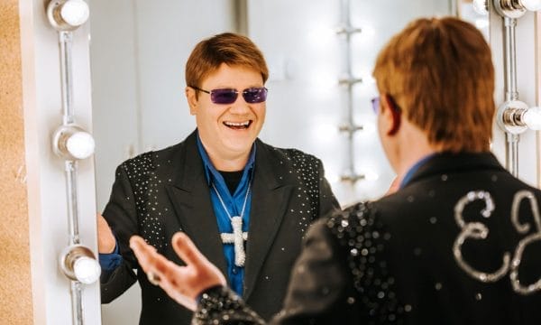 An Elton John lookalike smiles into the mirror, he is wearing a bedazzled black blazer, a silk blue shirt, and a large diamante crucifix necklace.