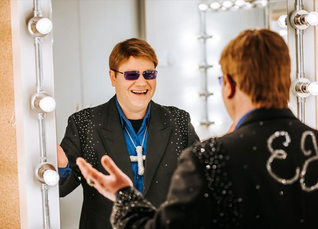 An Elton John lookalike smiles into the mirror, he is wearing a bedazzled black blazer, a silk blue shirt, and a large diamante crucifix necklace.