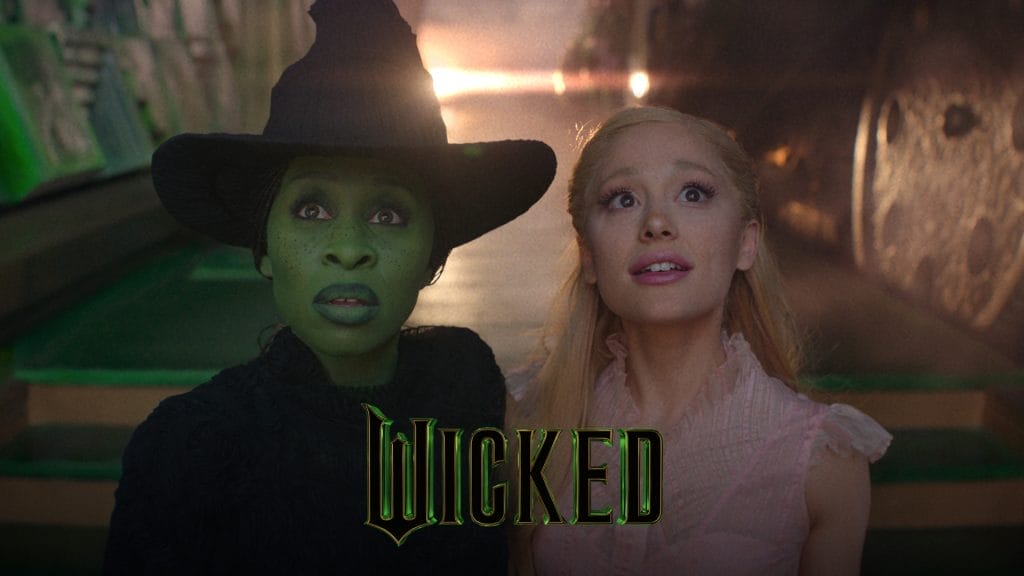 Ariana Grande and Cynthia Erivo both looking up in awe. Cynthia is wearing dark green facepaint and a pointed black hat, Ariana wears a long blonde wig and a pink dress. Text Reads: Wicked
