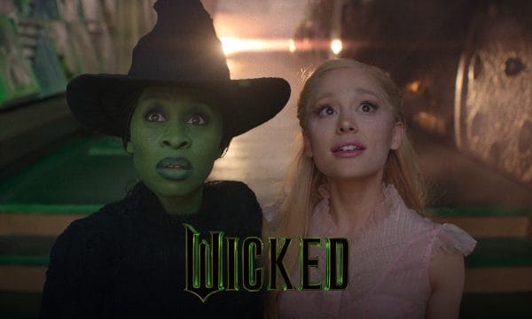 Ariana Grande and Cynthia Erivo both looking up in awe. Cynthia is wearing dark green facepaint and a pointed black hat, Ariana wears a long blonde wig and a pink dress. Text Reads: Wicked