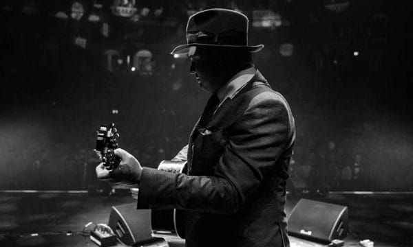 A black and white photo of Pete Doherty in side-profile. He is holding an acoustic guitar.