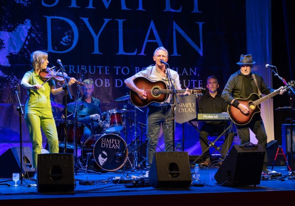 This image shows a live performance of a Bob Dylan tribute band called "Simply Dylan" on stage. The band consists of several musicians performing, including: A violinist wearing a bright lime green/yellow outfit A bassist in a patterned red/orange dress A drummer behind a kit with the "Simply Dylan" logo A lead guitarist/vocalist with an acoustic guitar Another guitarist in dark clothing with a hat The stage is lit with blue lighting and features a large "Simply Dylan" backdrop with text indicating it's a tribute to Bob Dylan. The setup includes microphones, monitors, and other standard stage equipment including what appears to be a Roland keyboard. The image captures the band mid-performance in what looks like a theatre or concert venue setting.