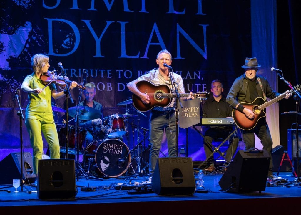 This image shows a live performance of a Bob Dylan tribute band called "Simply Dylan" on stage. The band consists of several musicians performing, including: A violinist wearing a bright lime green/yellow outfit A bassist in a patterned red/orange dress A drummer behind a kit with the "Simply Dylan" logo A lead guitarist/vocalist with an acoustic guitar Another guitarist in dark clothing with a hat The stage is lit with blue lighting and features a large "Simply Dylan" backdrop with text indicating it's a tribute to Bob Dylan. The setup includes microphones, monitors, and other standard stage equipment including what appears to be a Roland keyboard. The image captures the band mid-performance in what looks like a theatre or concert venue setting.