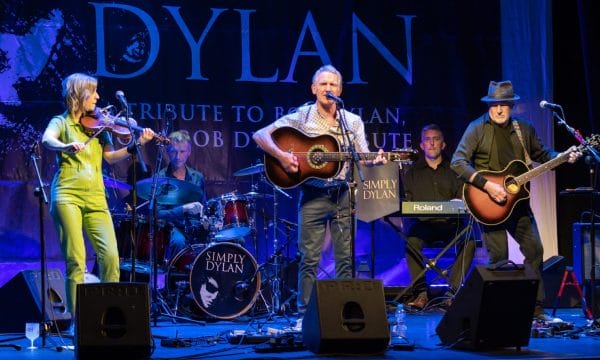 This image shows a live performance of a Bob Dylan tribute band called "Simply Dylan" on stage. The band consists of several musicians performing, including: A violinist wearing a bright lime green/yellow outfit A bassist in a patterned red/orange dress A drummer behind a kit with the "Simply Dylan" logo A lead guitarist/vocalist with an acoustic guitar Another guitarist in dark clothing with a hat The stage is lit with blue lighting and features a large "Simply Dylan" backdrop with text indicating it's a tribute to Bob Dylan. The setup includes microphones, monitors, and other standard stage equipment including what appears to be a Roland keyboard. The image captures the band mid-performance in what looks like a theatre or concert venue setting.