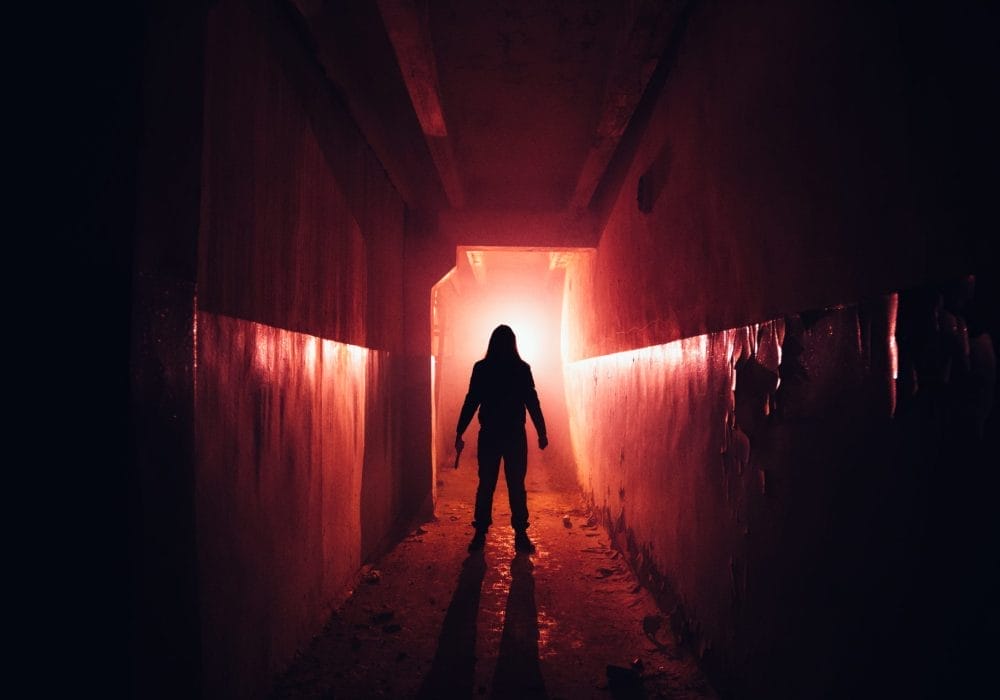 A silhouetted figure stands at the end of a dark, red-lit hallway. In one hand they are holding a weapon.