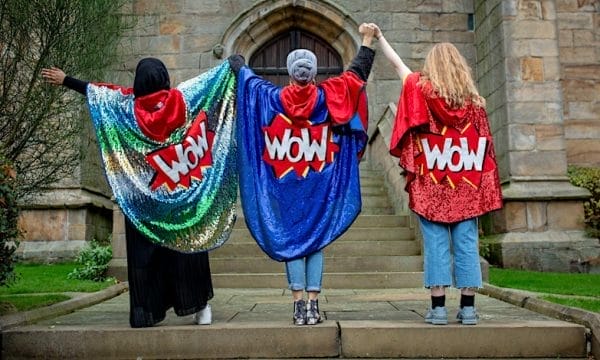 Three people wearing sparkling capes, all of the capes have a comic style WOW in the centre. They are standing holding hands, arms raised.