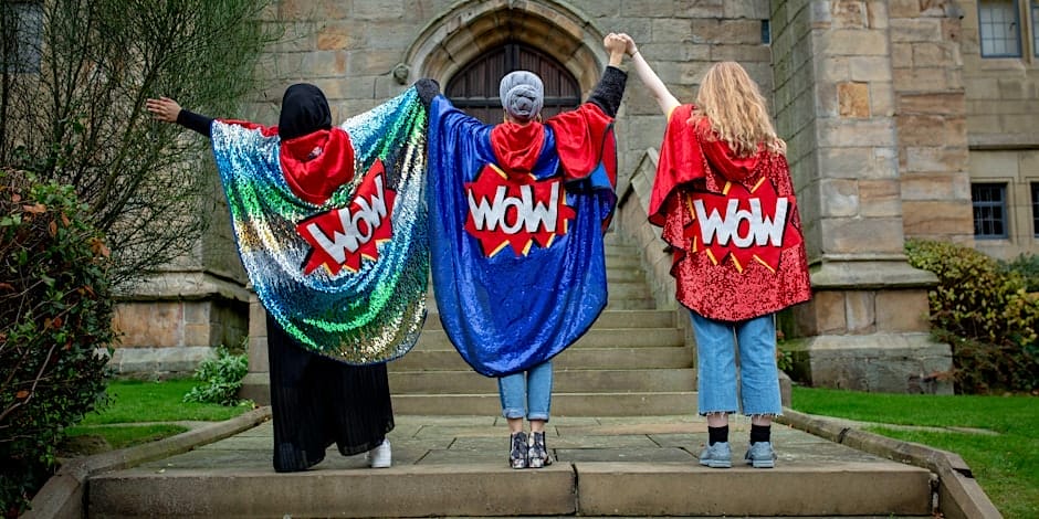 Three people wearing sparkling capes, all of the capes have a comic style WOW in the centre. They are standing holding hands, arms raised.
