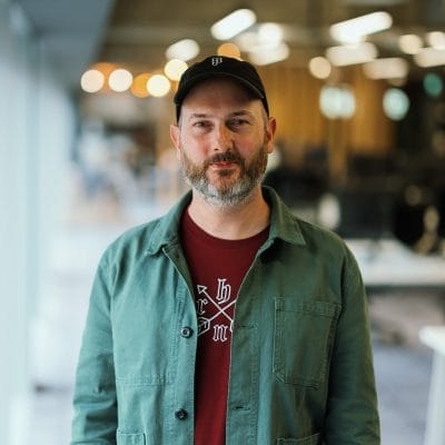 Headshot of Phil Saunders. He is wearing a black baseball cap, and a green jacket over a dark red t-shirt.