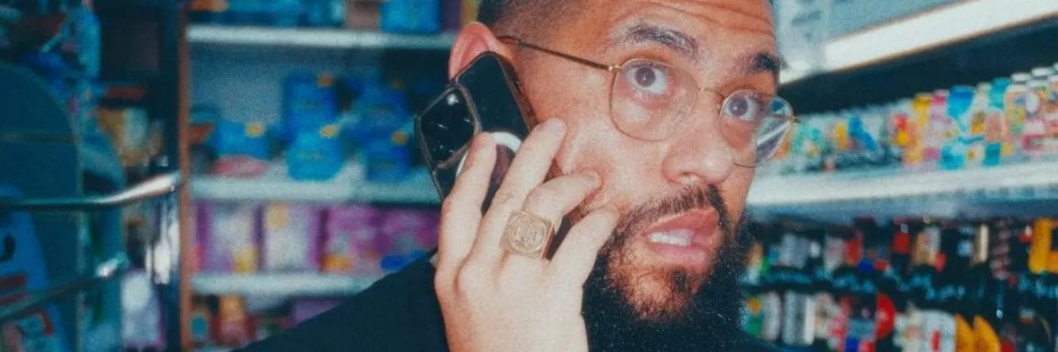 Jamali in what appears to be a convenience store or small shop. The person is wearing a black shirt and has glasses and a beard. They're talking on a phone while holding what looks like beverage bottles in their other hand. They have visible tattoos on their arm and are wearing a ring. The photo appears to be taken with a vintage or filtered effect, giving it a slightly blue-tinted color palette. The background shows store shelves stocked with various drinks and products.