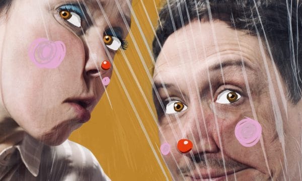 two people behind a plastic or cellophane-like transparent material, giving a wrapped or enclosed effect. The background is a vibrant yellow-orange color. Both individuals have digitally added elements on their faces - they have cartoon-like eyes with exaggerated white areas around amber/orange irises, and pink circular spots on their cheeks and red circular spots on their noses, resembling clown makeup or digital stickers.