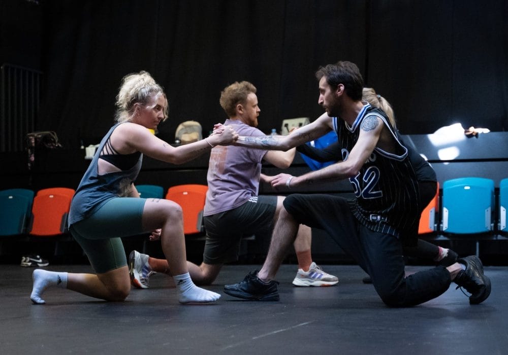 A group of people in a flat-floor theatre space. Two people in the foreground are down on one knee facing each other, they are clutching hands and looking intently at each other.