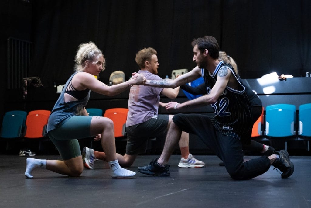 A group of people in a flat-floor theatre space. Two people in the foreground are down on one knee facing each other, they are clutching hands and looking intently at each other.