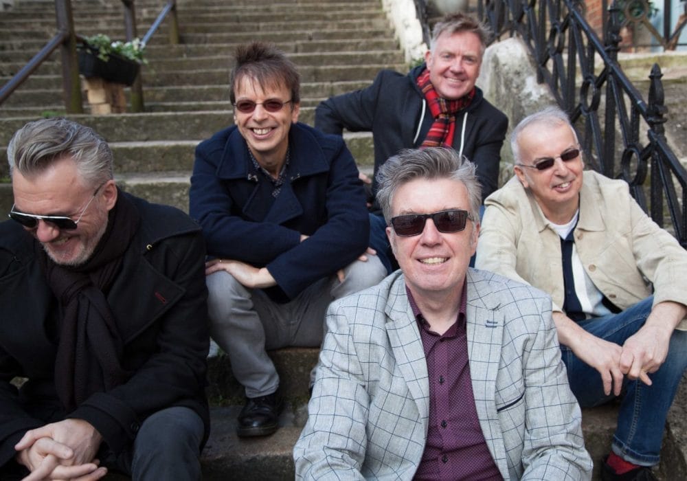 The Undertones, a group of five older white men, are sitting on some stone steps. They are all smiling and laughing.