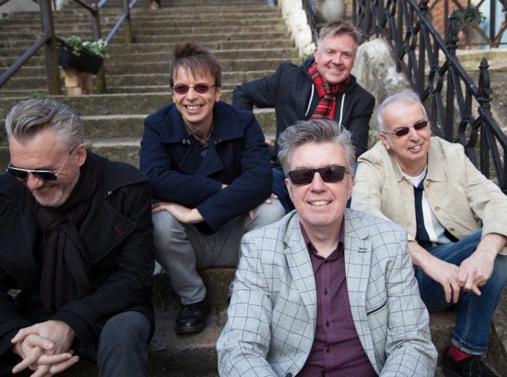 The Undertones, a group of five older white men, are sitting on some stone steps. They are all smiling and laughing.