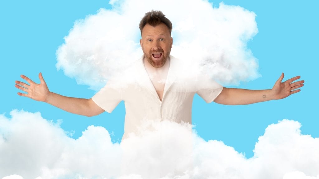 Comedian Jason Byrne, a middle-aged white man, wears a bright white shirt, and stands with his arms outstretched, grinning. His head is surrounded by fluffy white clouds.