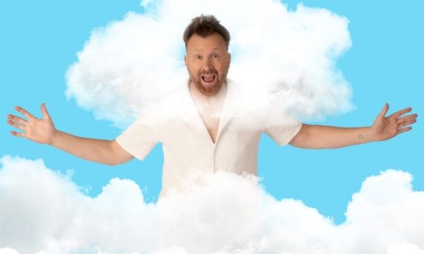 Comedian Jason Byrne, a middle-aged white man, wears a bright white shirt, and stands with his arms outstretched, grinning. His head is surrounded by fluffy white clouds.