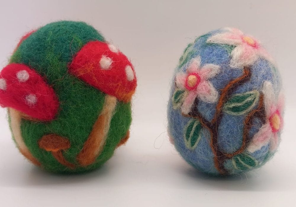 A pair of decorated needle felt eggs, one is green and decorated with red toadstools, and the other is light blue decorated with cherry blossoms.