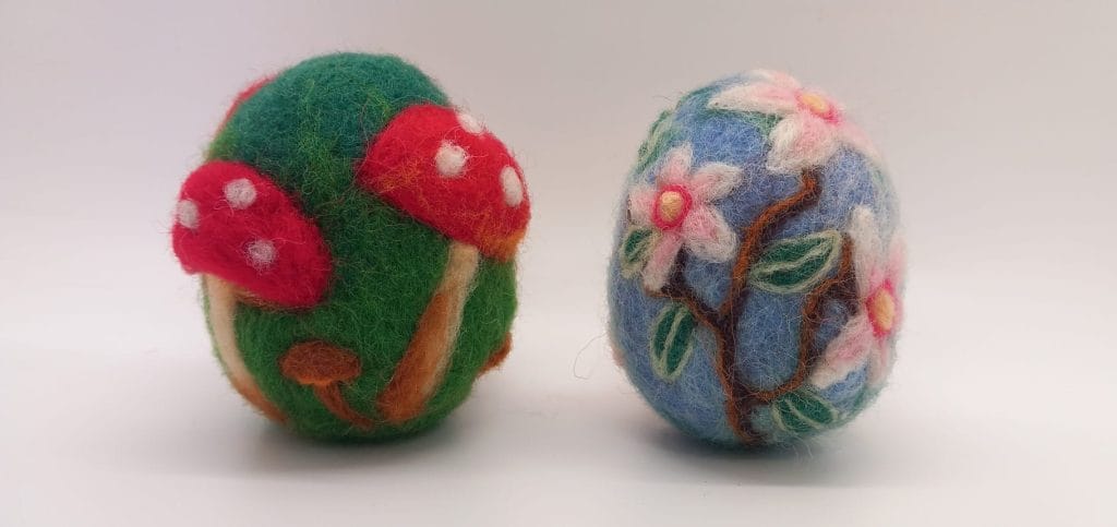 A pair of decorated needle felt eggs, one is green and decorated with red toadstools, and the other is light blue decorated with cherry blossoms.