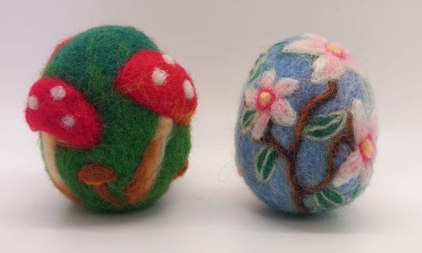 A pair of decorated needle felt eggs, one is green and decorated with red toadstools, and the other is light blue decorated with cherry blossoms.