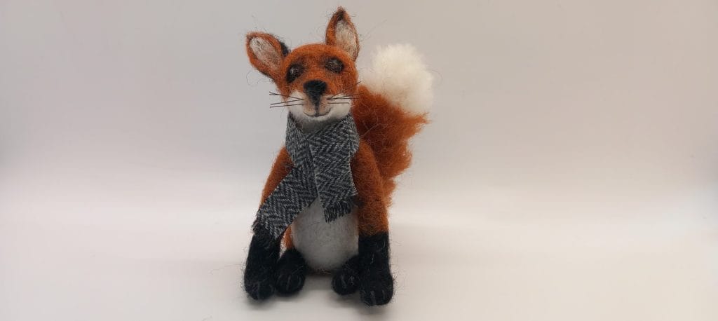 A needle felt fox, wearing a grey scarf.