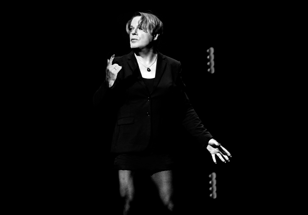 Comedian Eddie Izzard, a white person with medium length hair, stands against a black backdrop. They are wearing a black jacket, and a black necklace. The image is black and white.