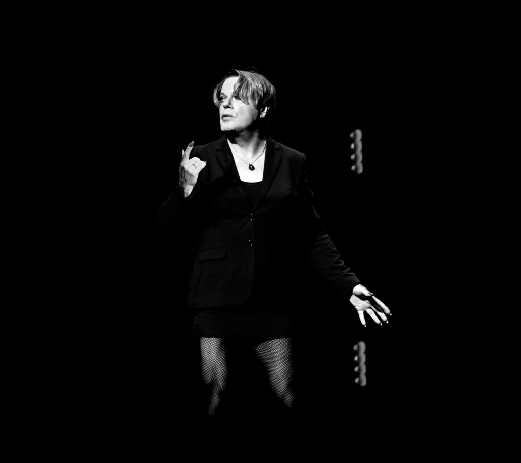 Comedian Eddie Izzard, a white person with medium length hair, stands against a black backdrop. They are wearing a black jacket, and a black necklace. The image is black and white.