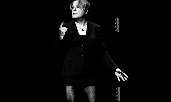 Comedian Eddie Izzard, a white person with medium length hair, stands against a black backdrop. They are wearing a black jacket, and a black necklace. The image is black and white.