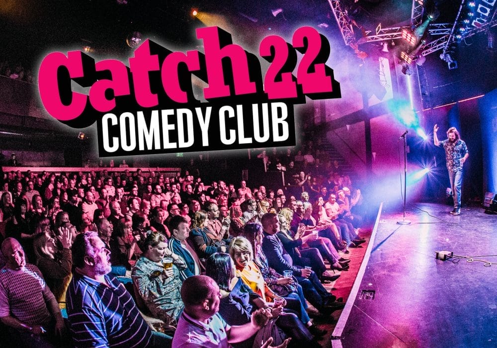 A promotional photo for Catch 22 Comedy Club. The vibrant scene depicts a crowded comedy venue with a large audience seated in front of a stage. On stage, a performer (likely a comedian) can be seen standing with a microphone. The venue is lit with theatrical lighting in purple and blue hues, creating an energetic atmosphere.